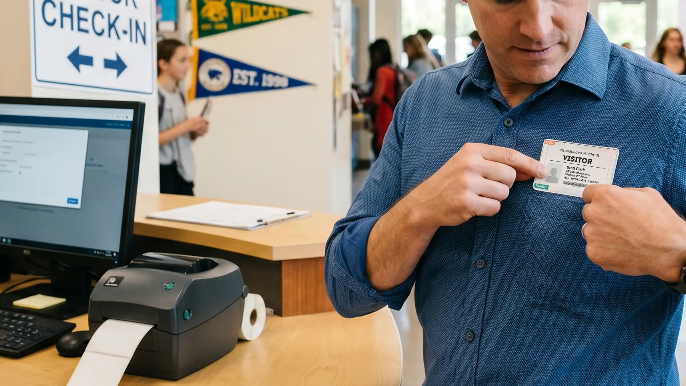 A visitor at a school front desk putting on a color-coded visitor badge beside a badge printer